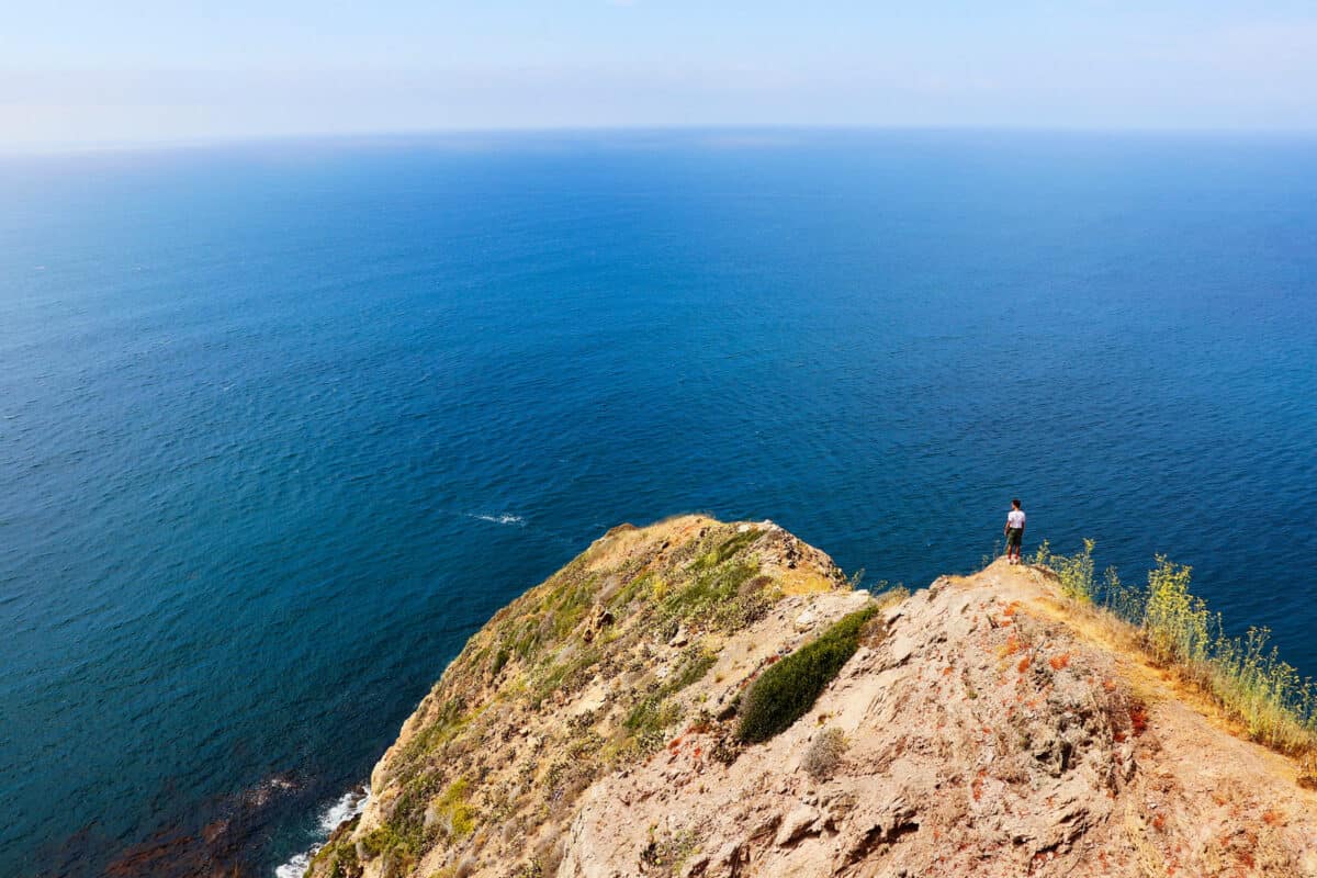 The view from Arrow Point: A rocky outcrop in the foreground with a person standing at the edge, overlooking a vast ocean and blue sky.